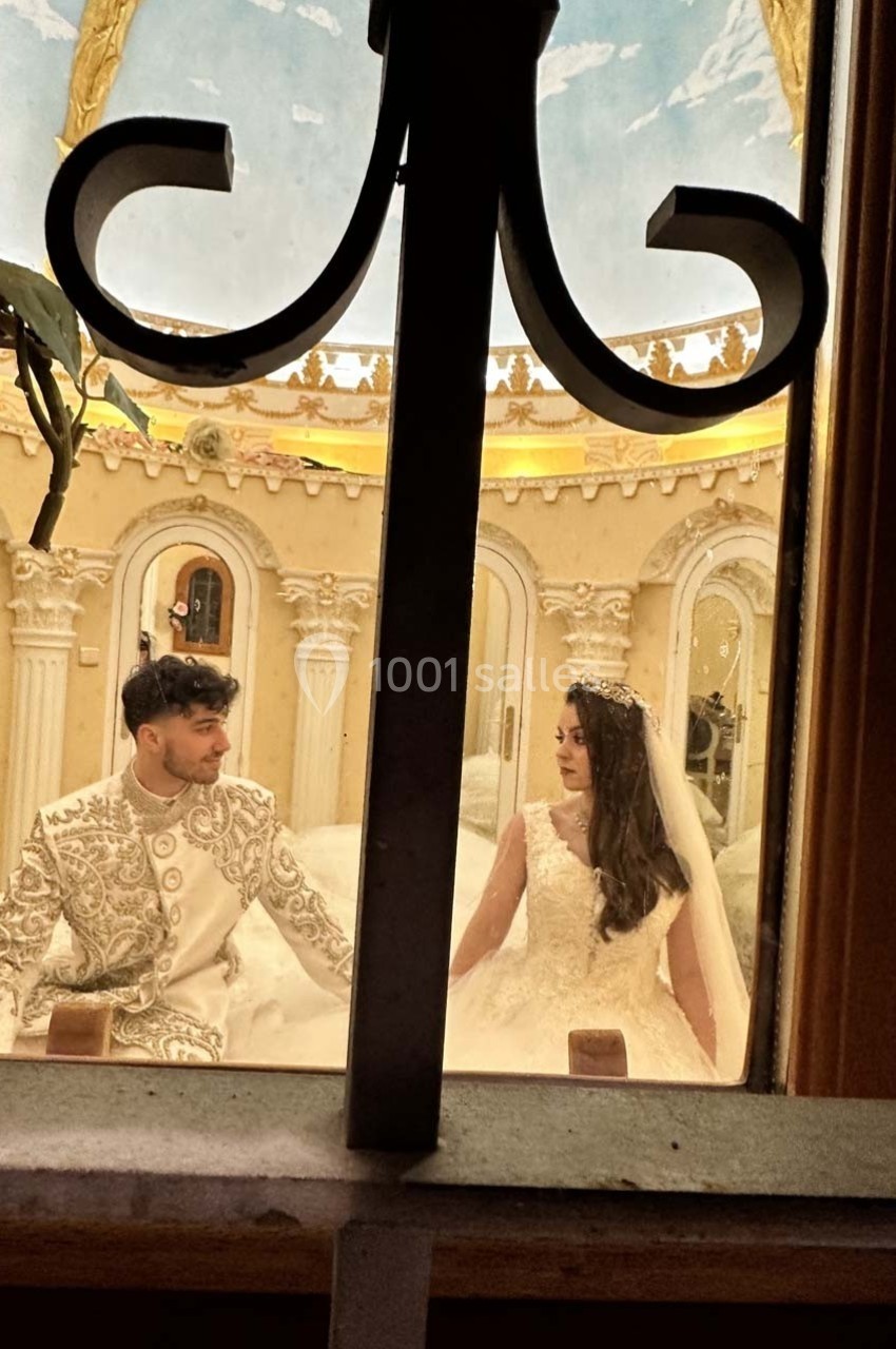 Un couple en tenue de mariage assis dans un décor intérieur élégant vu à travers une fenêtre avec une grille.