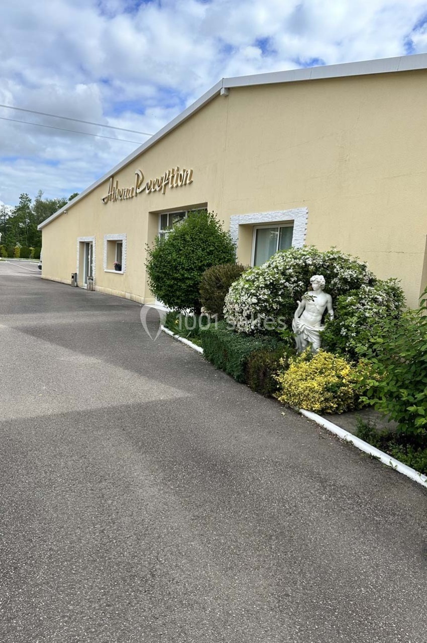 Bâtiment beige avec enseigne ’Auberge Réception’, bordé de buissons, statue décorative et allée asphaltée.