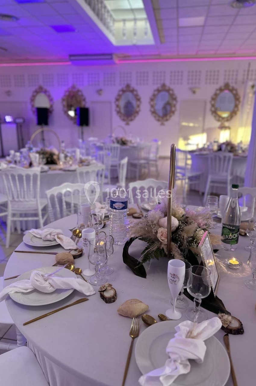 Salle de réception décorée avec des tables rondes, nappes blanches, centres de table floraux et éclairage tamisé.
