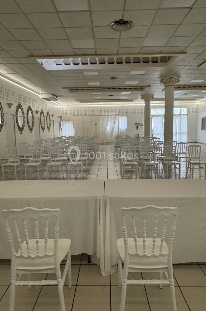 Salle de réception lumineuse avec chaises blanches alignées, tables recouvertes de nappes et miroirs décoratifs au mur.