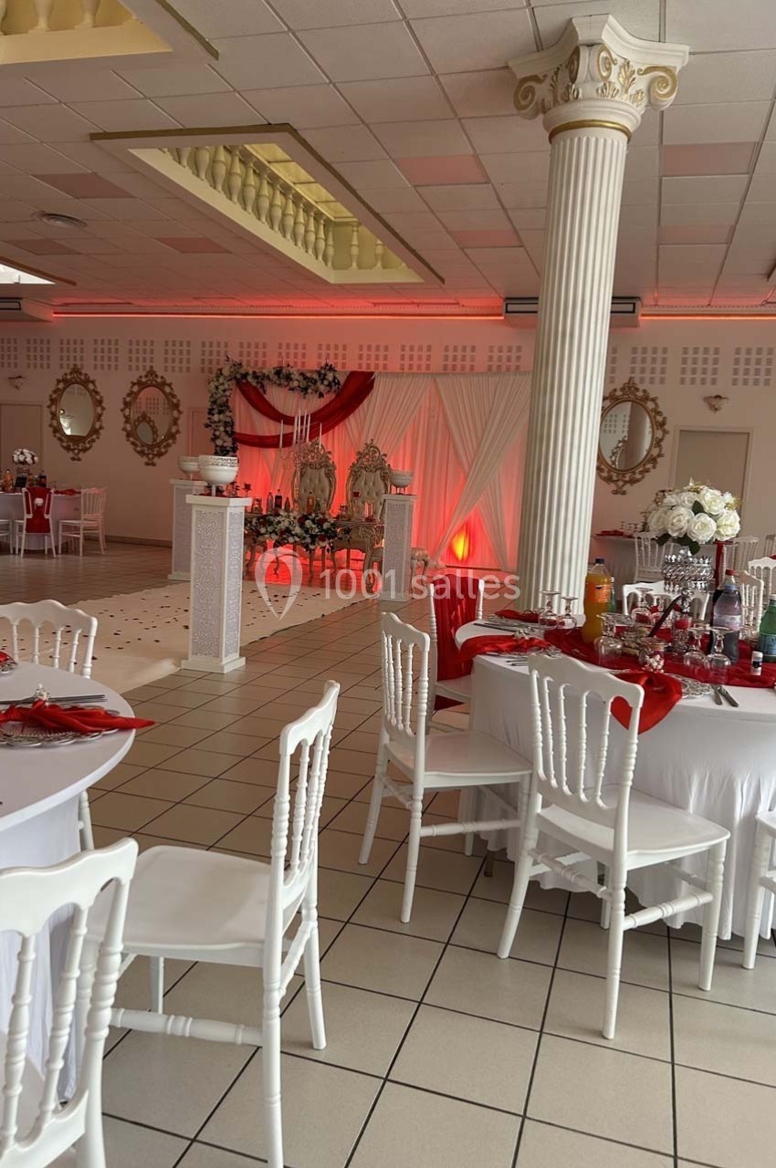 Salle de réception décorée avec des tables blanches, des nappes rouges et un espace central avec des fauteuils ornés.
