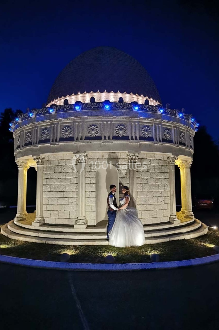 Un couple en tenue de mariage pose devant un pavillon éclairé de nuit, entouré d'un décor sombre.