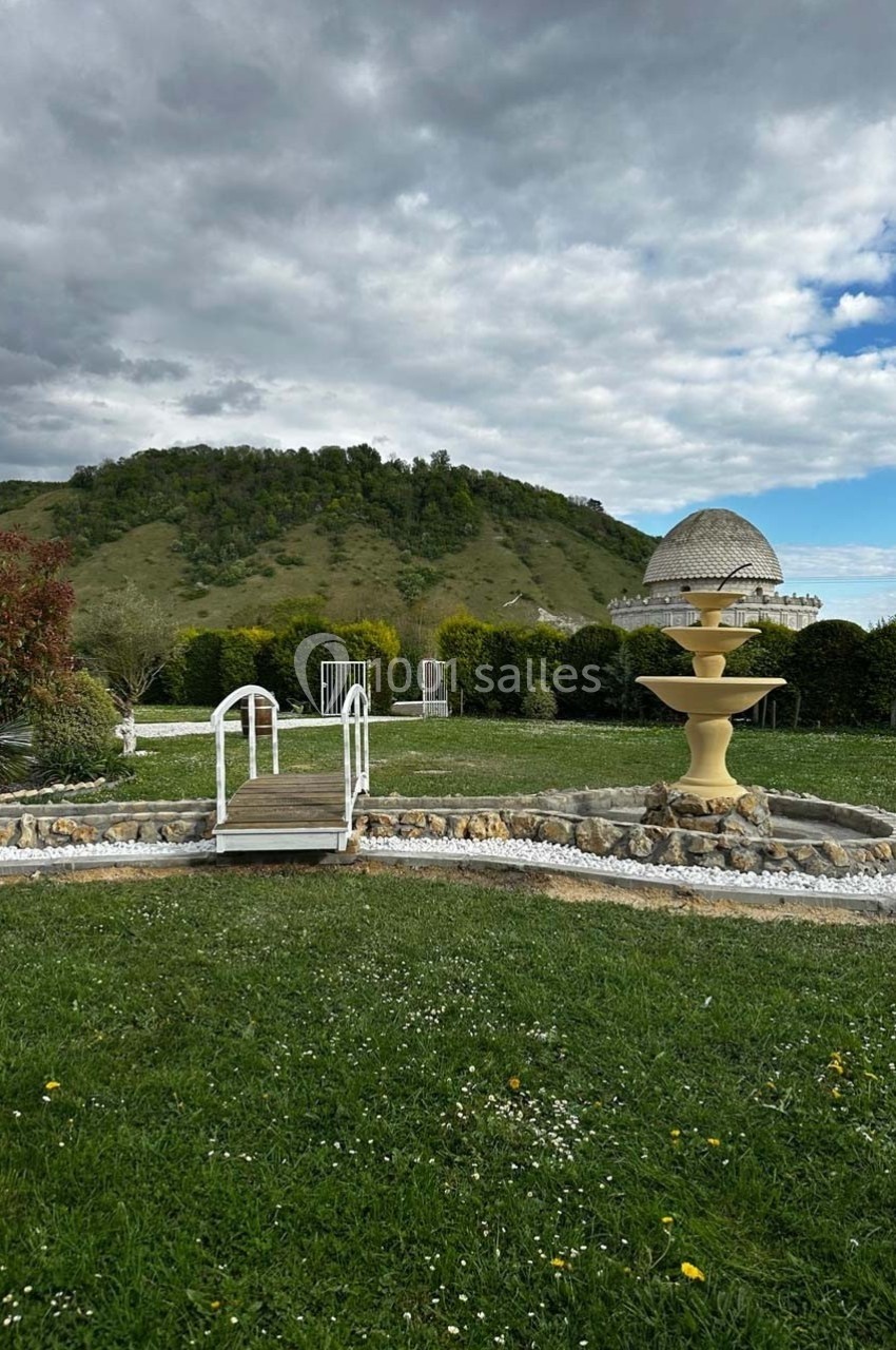 Fontaine beige entourée de pelouse, petit pont en bois et colline verdoyante sous un ciel nuageux.