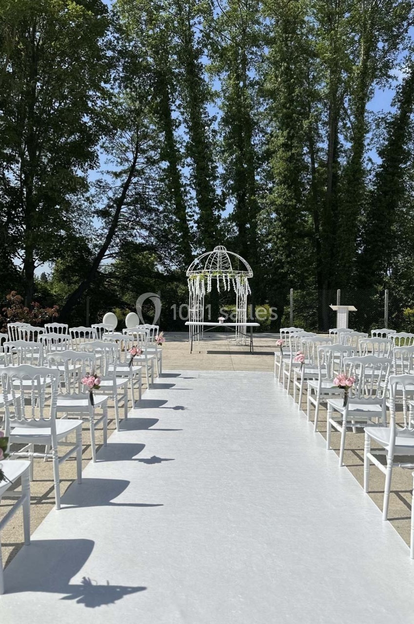 Allée centrale bordée de chaises blanches décorées de fleurs roses, menant à une arche de mariage sous des arbres.