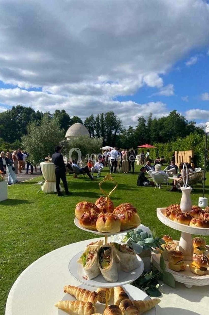Buffet en plein air avec viennoiseries et wraps, invités rassemblés dans un jardin sous un ciel partiellement nuageux.