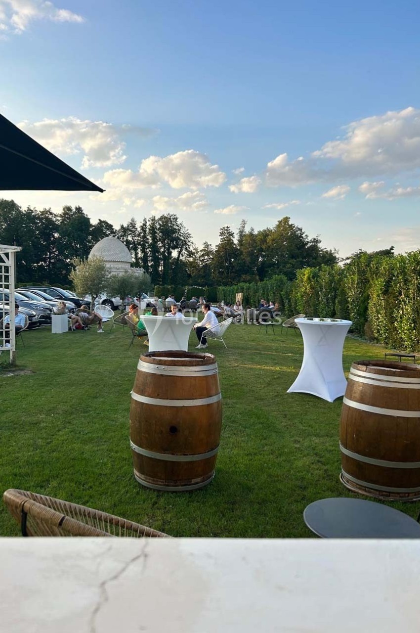 Espace extérieur avec tables hautes, tonneaux en bois et invités assis sur une pelouse, sous un ciel dégagé.