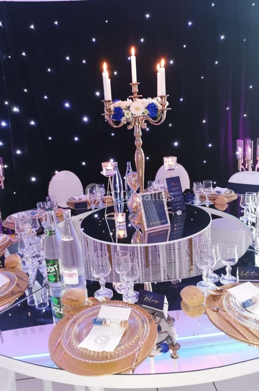 Table élégamment dressée avec chandeliers, vaisselle dorée, verres et décor lumineux en arrière-plan.