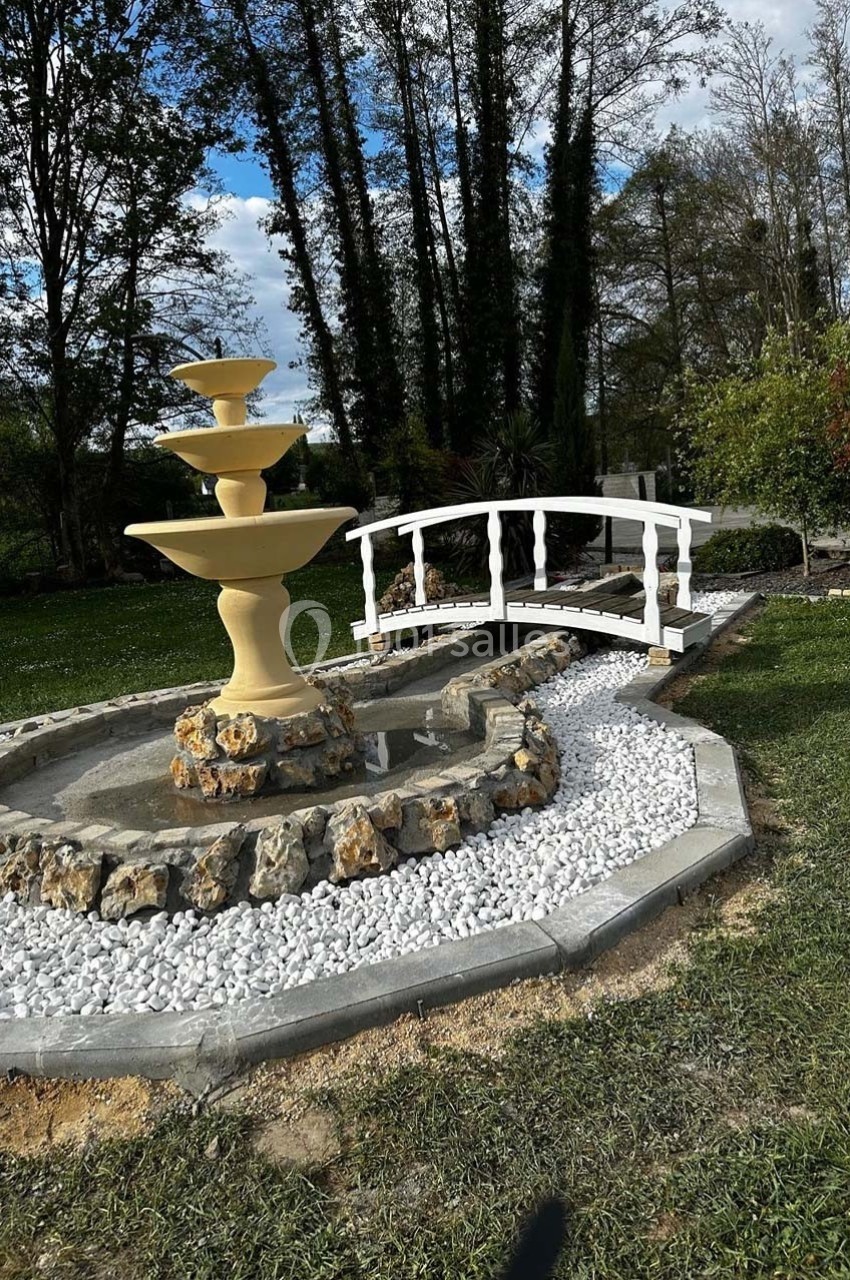 Fontaine beige à trois niveaux entourée de galets blancs, avec un petit pont en bois blanc dans un jardin verdoyant.