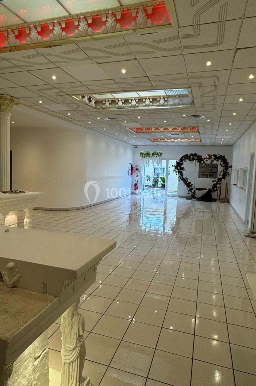 Salle lumineuse avec carrelage blanc, colonnes décoratives et arche en forme de cœur ornée de fleurs au fond.