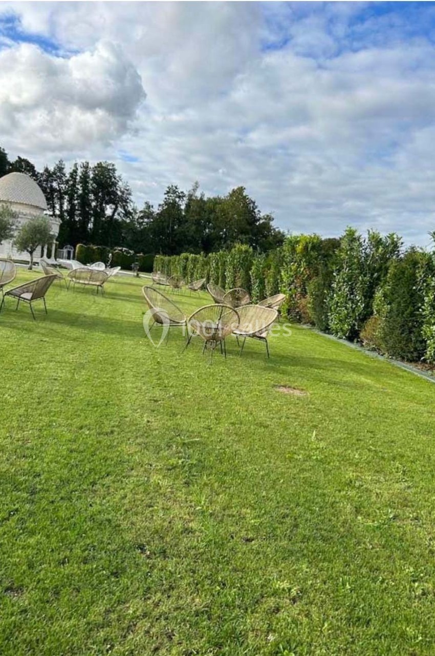 Chaises en osier disposées sur une pelouse verte, entourées de haies et d'arbres sous un ciel partiellement nuageux.