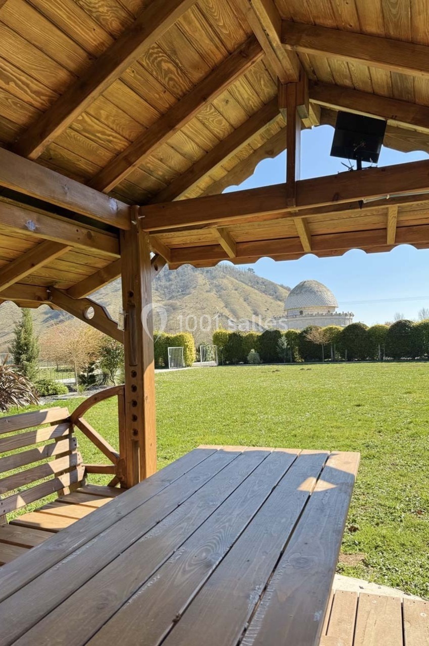 Vue depuis un kiosque en bois sur une table, un banc, une pelouse, des arbres et des collines en arrière-plan.