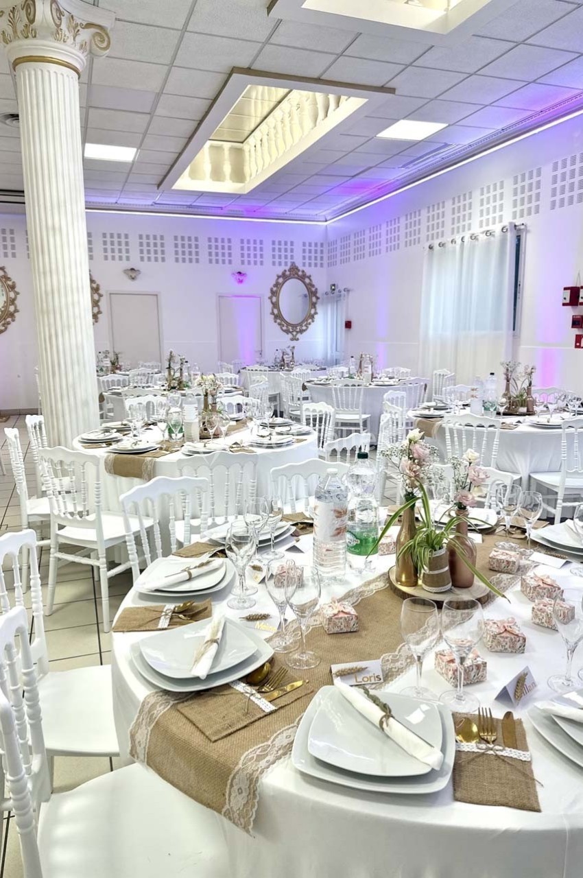 Salle de réception décorée avec des tables rondes dressées, nappes blanches, chemins de table en toile de jute et vaisselle…