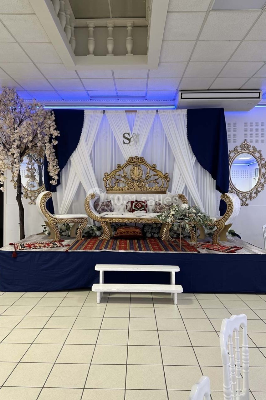 Décor de scène avec canapé doré, rideaux blancs et bleus, tapis rouge, fleurs artificielles et miroirs muraux.