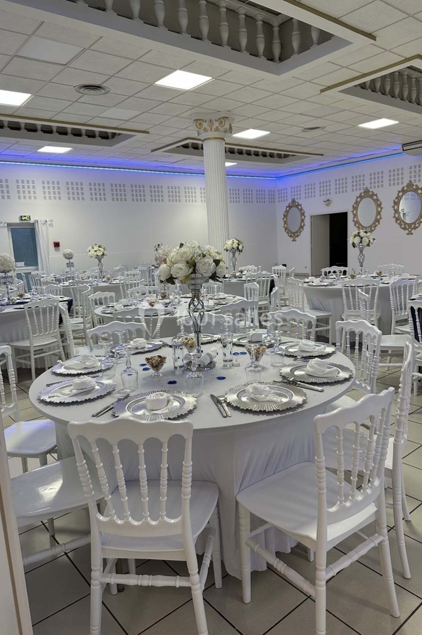 Salle de réception décorée avec des tables rondes dressées, chaises blanches et centre de table fleuris.