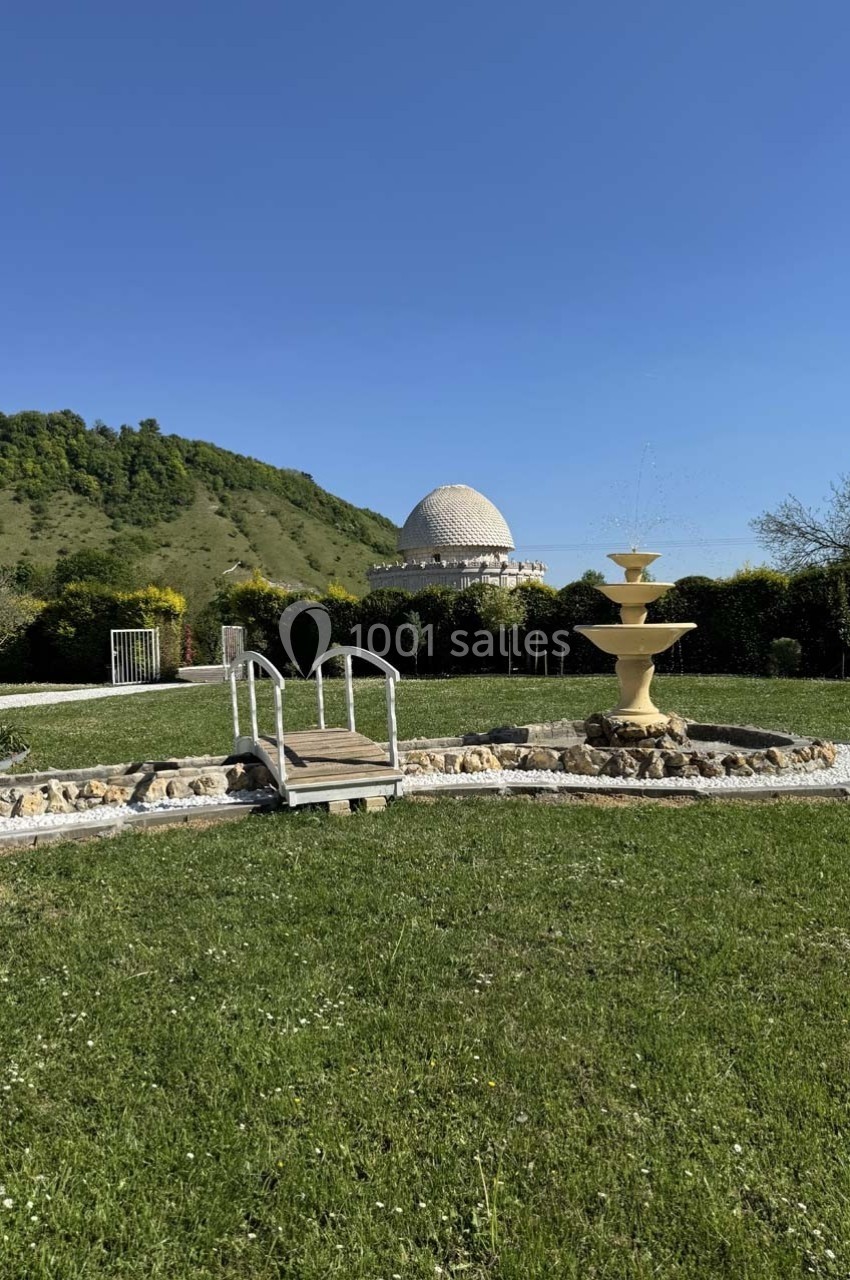 Petit pont en bois traversant un ruisseau artificiel, avec une fontaine et un dôme blanc en arrière-plan, entourés de…
