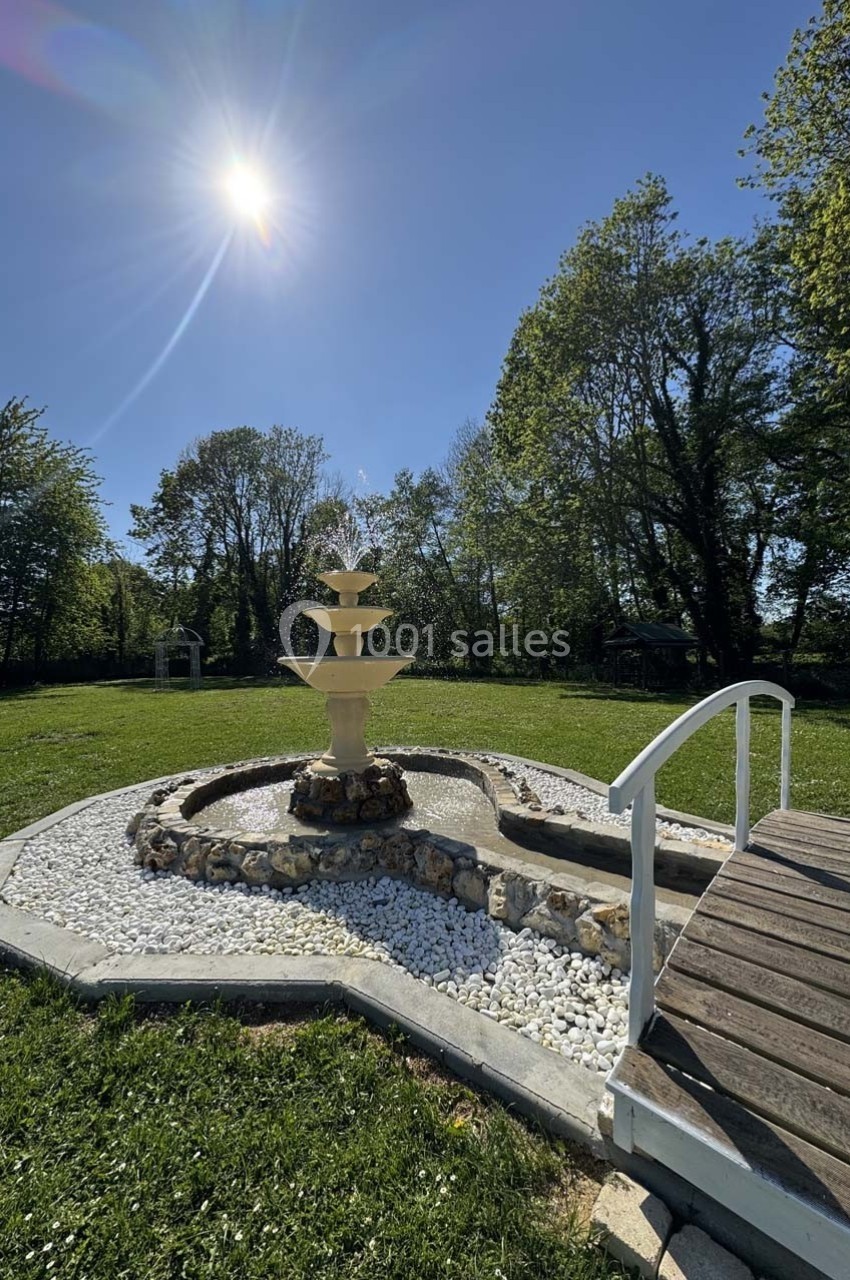 Fontaine en pierre entourée de galets blancs, située dans un jardin verdoyant sous un ciel ensoleillé.