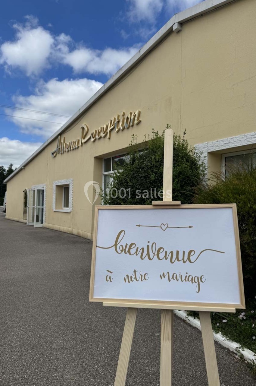 Panneau ’Bienvenue à notre mariage’ sur un chevalet devant un bâtiment beige par temps ensoleillé.