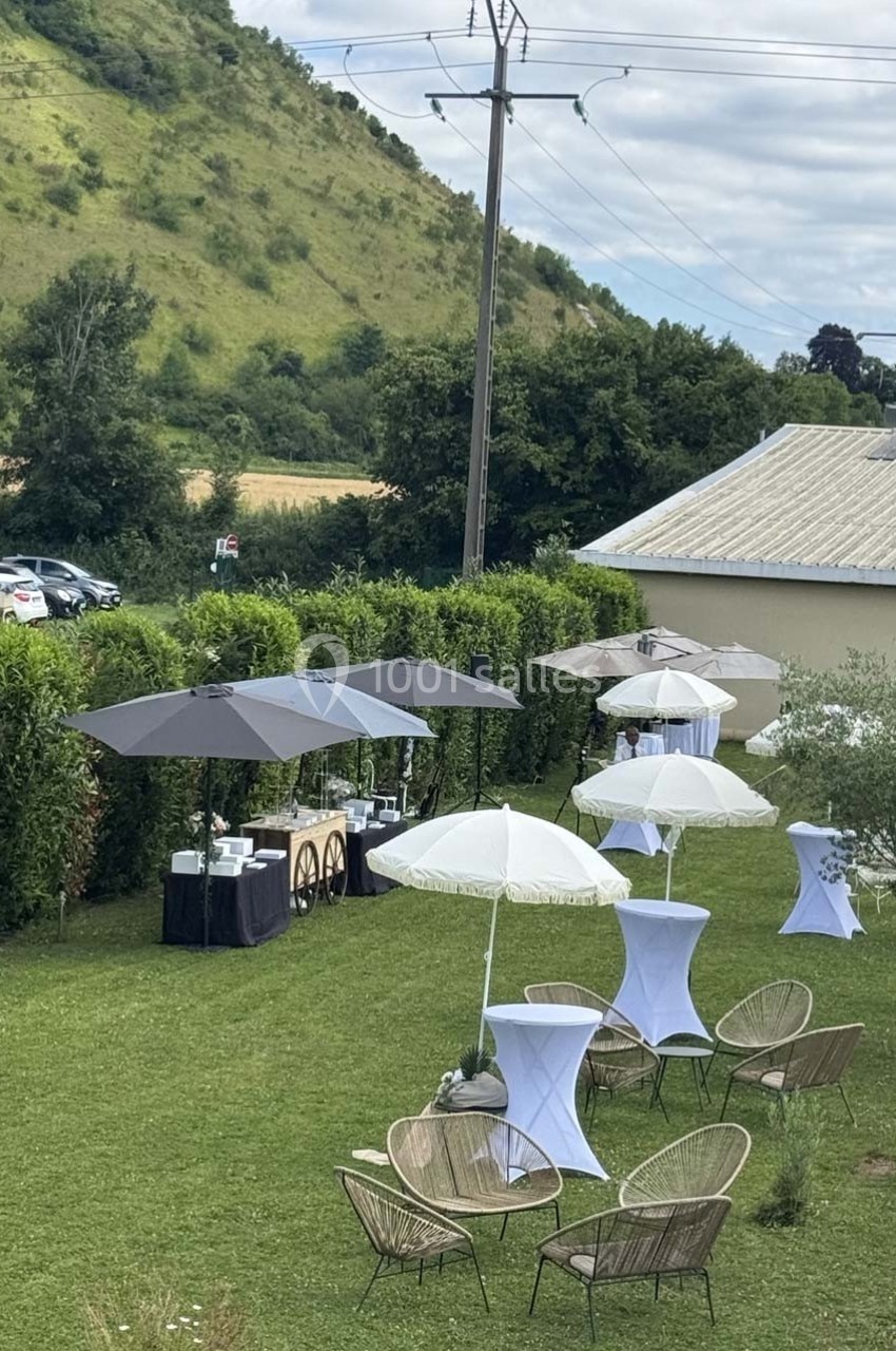 Espace extérieur aménagé avec chaises, tables hautes et parasols, entouré de verdure et d'une colline en arrière-plan.