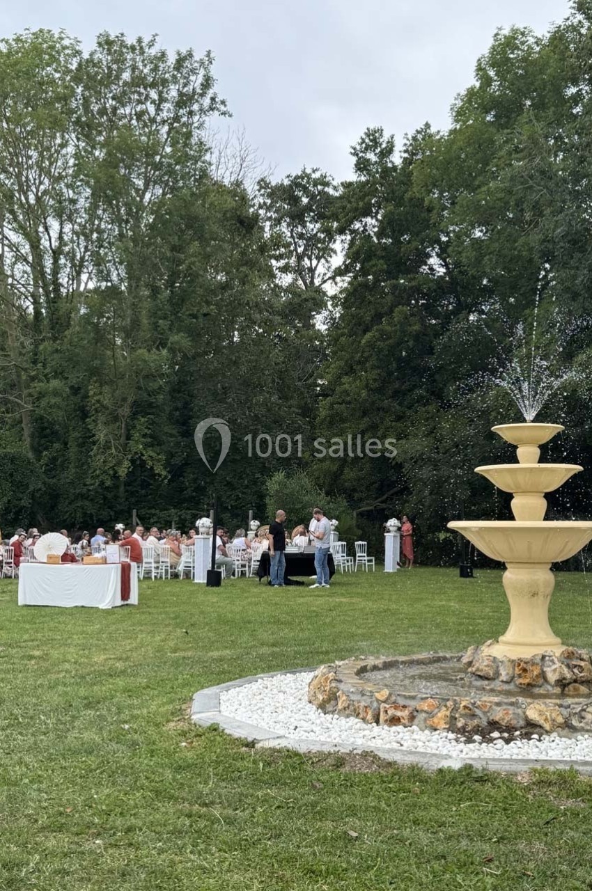 Fontaine beige à plusieurs niveaux au premier plan, avec des invités rassemblés autour de tables dans un jardin verdoyant.