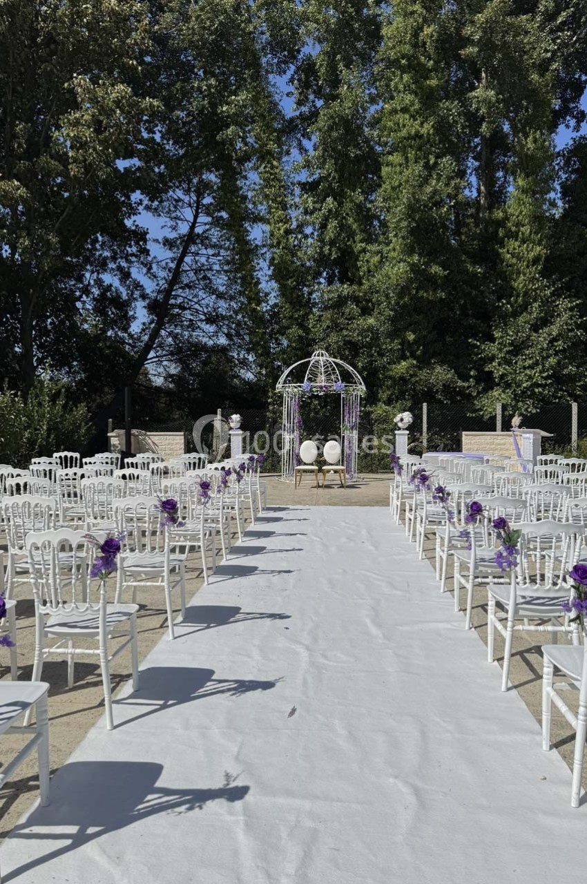 Allée centrale bordée de chaises blanches décorées de fleurs violettes menant à une arche de mariage en extérieur.
