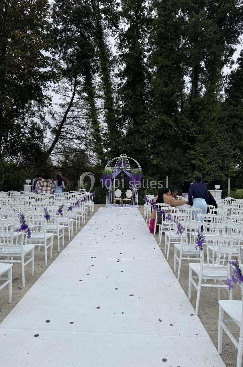 Allée centrale bordée de chaises blanches décorées de fleurs violettes, menant à une arche ornée de végétation.