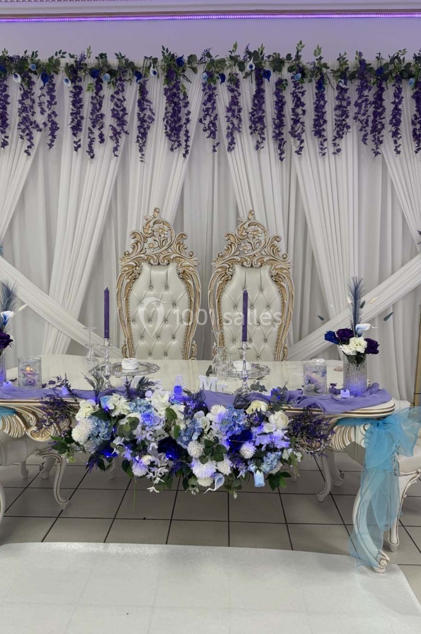 Décoration de table de mariage avec chaises ornées, fleurs blanches et violettes, et rideaux blancs décorés de guirlandes.