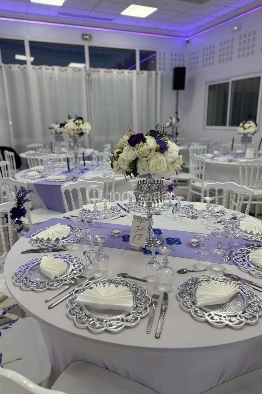 Salle de réception décorée avec des tables rondes, nappes violettes, vaisselle élégante et centres de table floraux.
