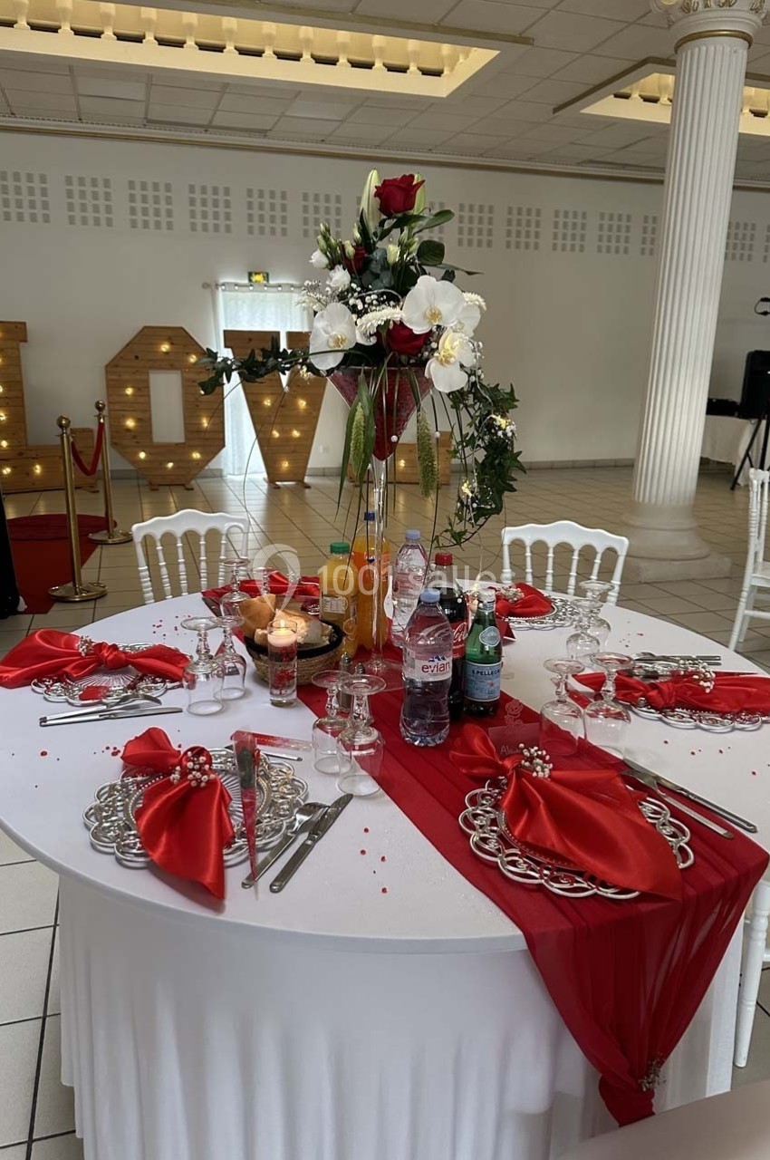 Table de réception élégamment décorée avec nappes blanches, chemins de table rouges, fleurs et boissons variées.