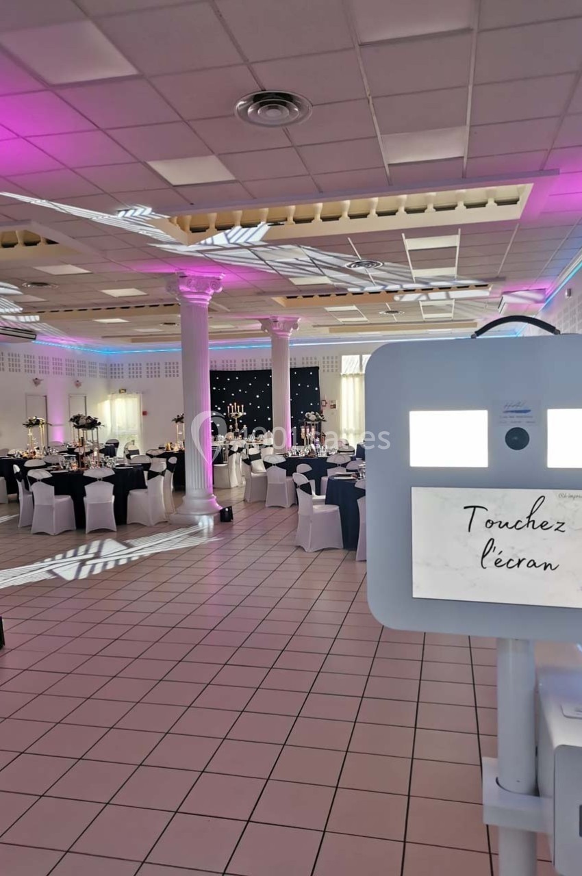 Salle de réception décorée avec des tables dressées et un photobooth en premier plan sous un éclairage rose.