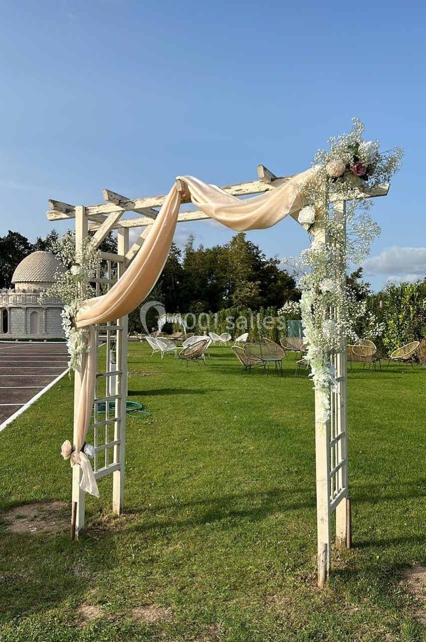 Arche en bois décorée de fleurs blanches et de tissu beige, installée sur une pelouse sous un ciel dégagé.