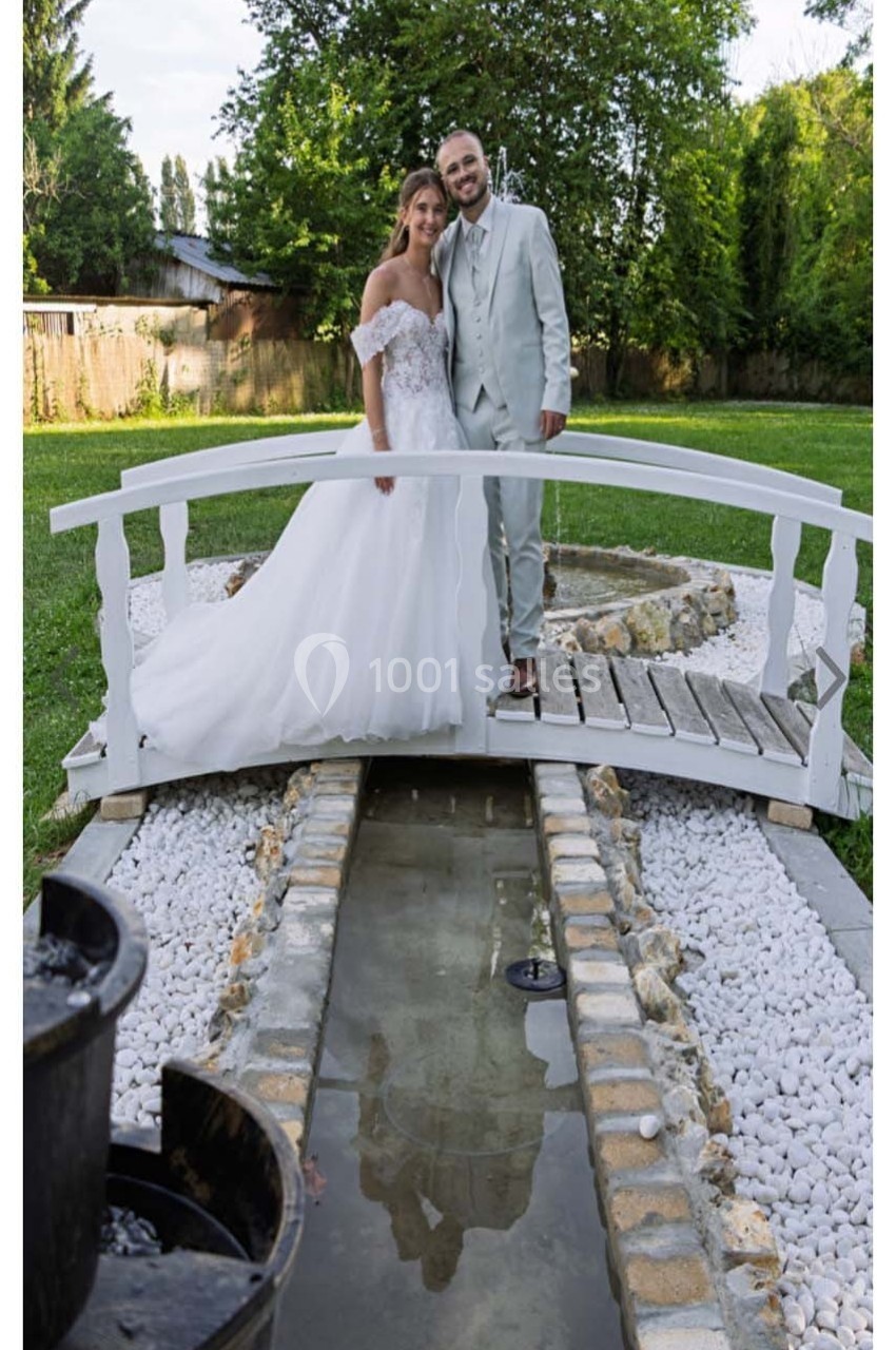 Un couple en tenue de mariage pose sur un petit pont en bois au-dessus d'un ruisseau dans un jardin verdoyant.
