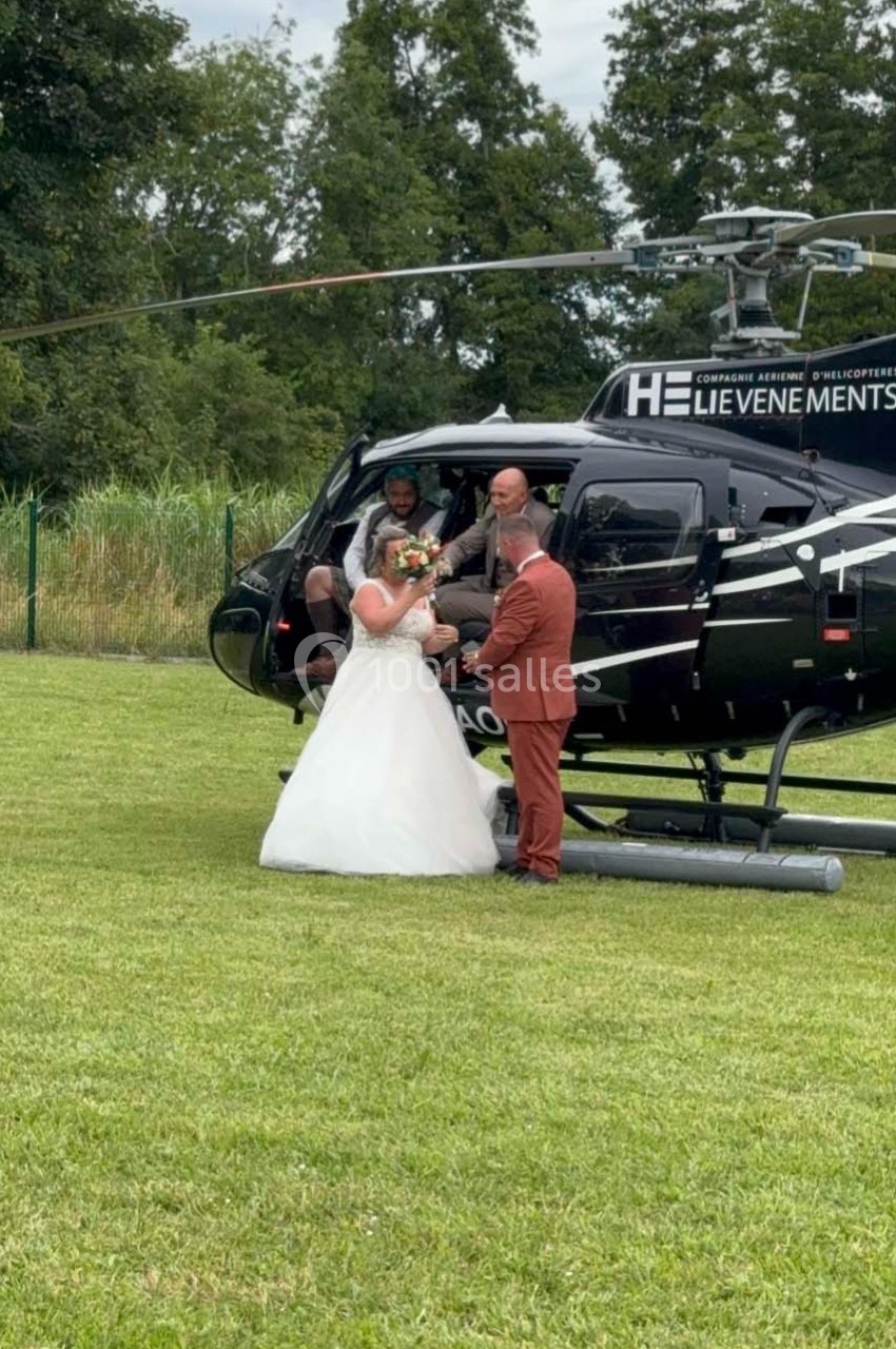 Un couple en tenue de mariage descend d'un hélicoptère noir posé sur une pelouse, entouré de verdure.