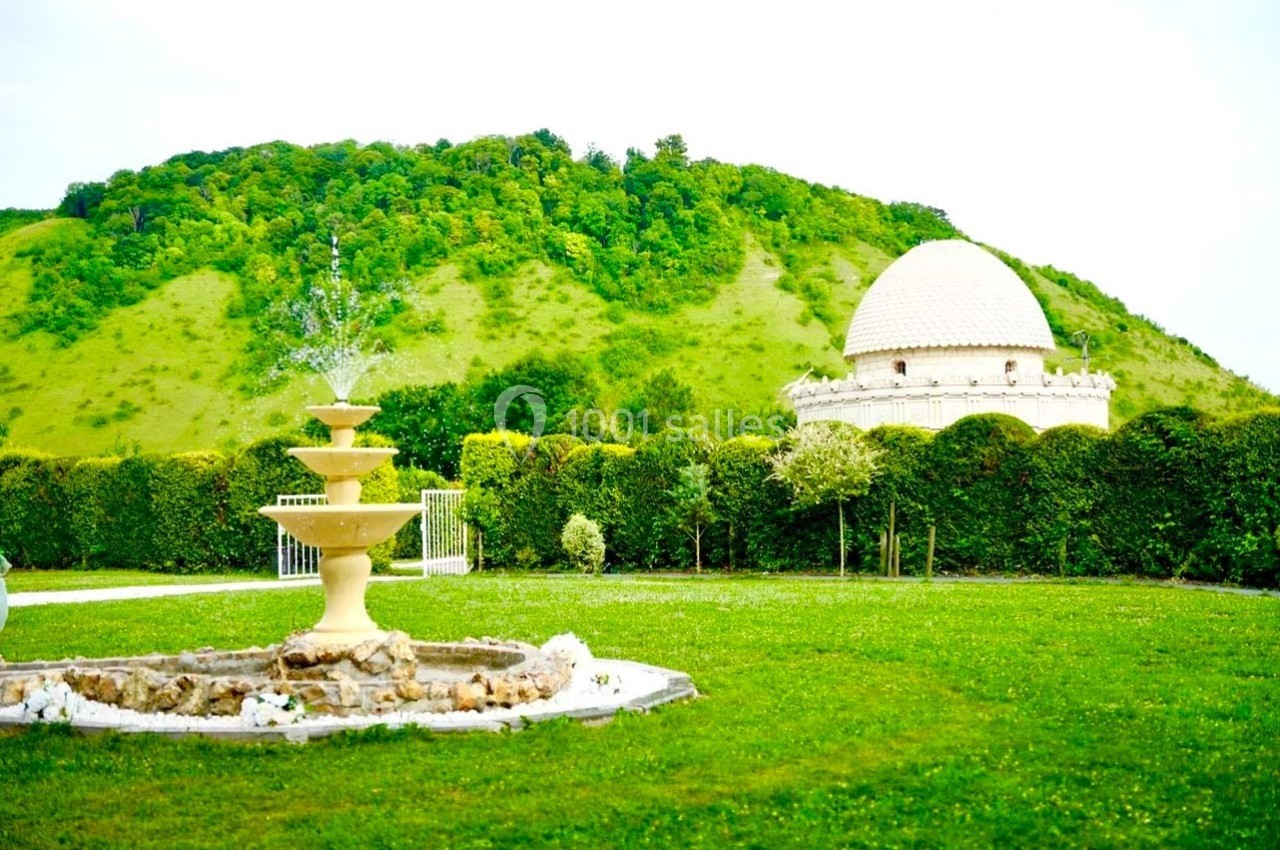 Fontaine décorative dans un jardin verdoyant avec une colline boisée et un bâtiment à dôme en arrière-plan.