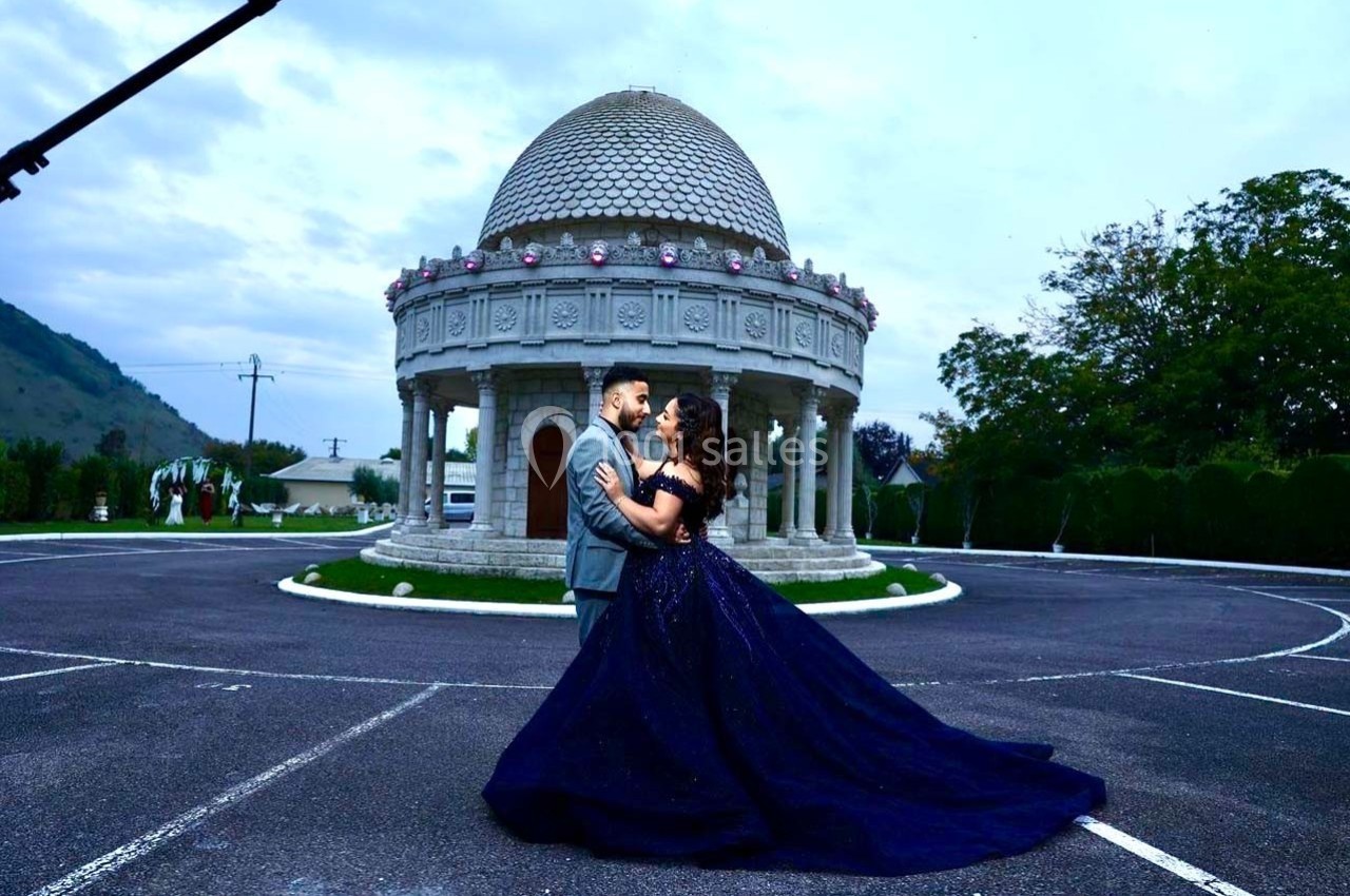 Un couple élégant danse devant un kiosque en pierre dans un espace extérieur pavé, sous un ciel nuageux.