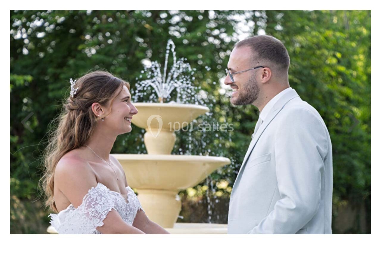 Un couple en tenue de mariage se regarde en souriant devant une fontaine dans un jardin verdoyant.