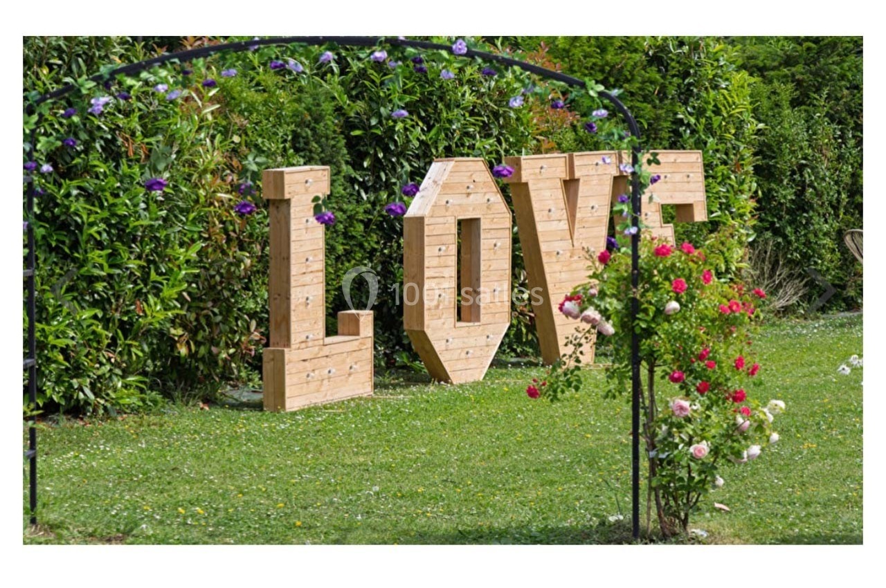 Lettres en bois formant le mot ’LOVE’ dans un jardin verdoyant, entourées de fleurs et d'une arche métallique.