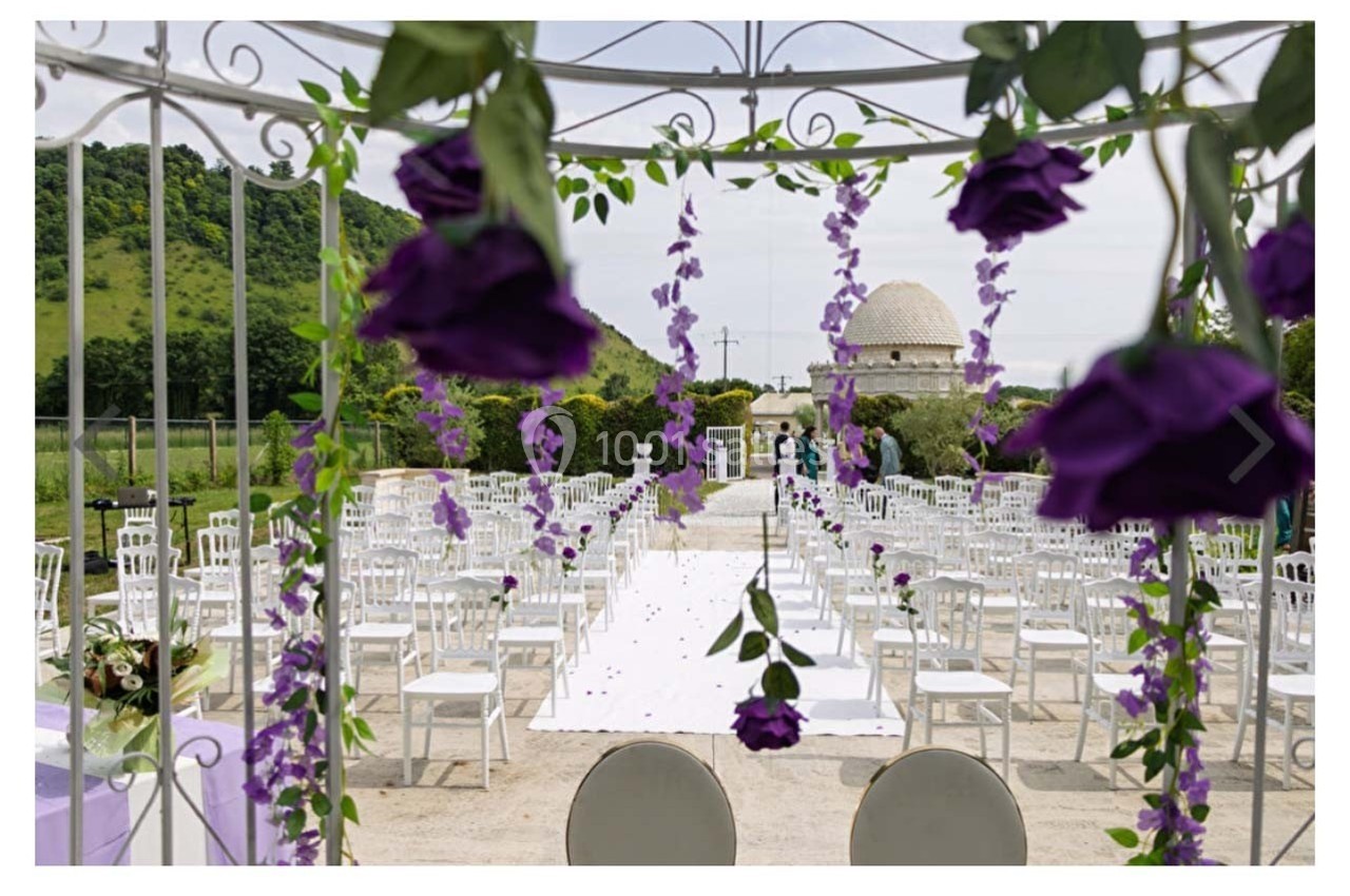 Allée centrale décorée de fleurs violettes menant à un espace de cérémonie en plein air avec des chaises blanches alignées.