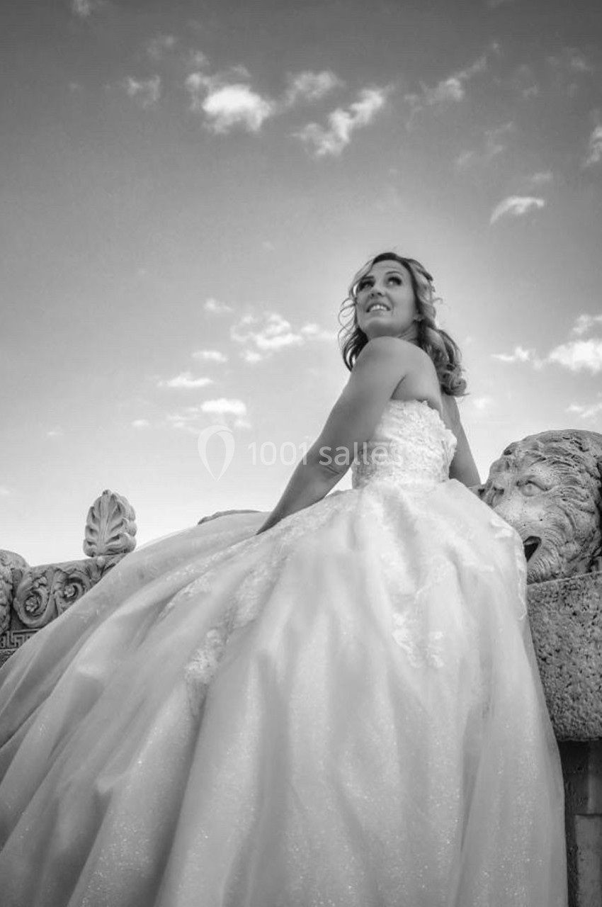 Femme en robe de mariée assise sur un muret décoré de sculptures, regardant vers le ciel partiellement nuageux.