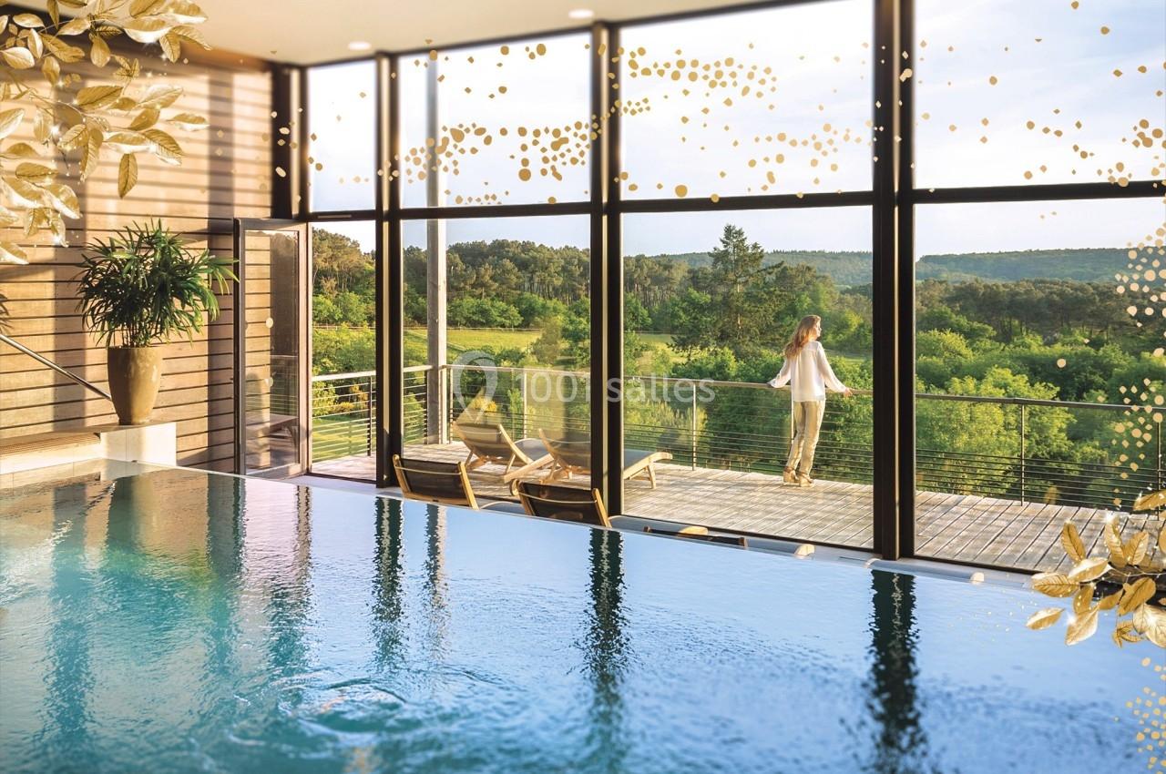 Vue intérieure d'une piscine avec baies vitrées donnant sur une terrasse en bois et un paysage verdoyant.