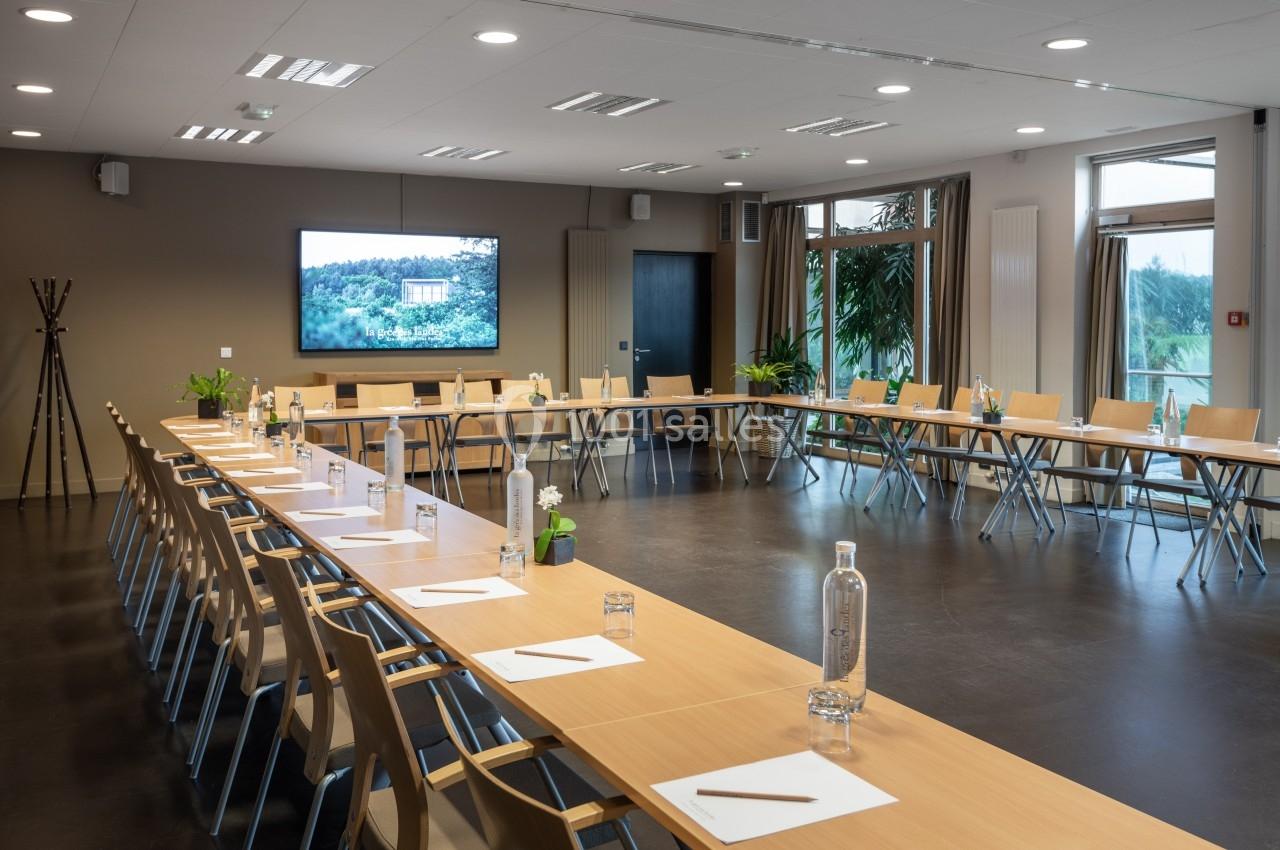 Salle de réunion lumineuse avec tables en U, chaises en bois, bouteilles d'eau, blocs-notes et écran affichant un paysage.