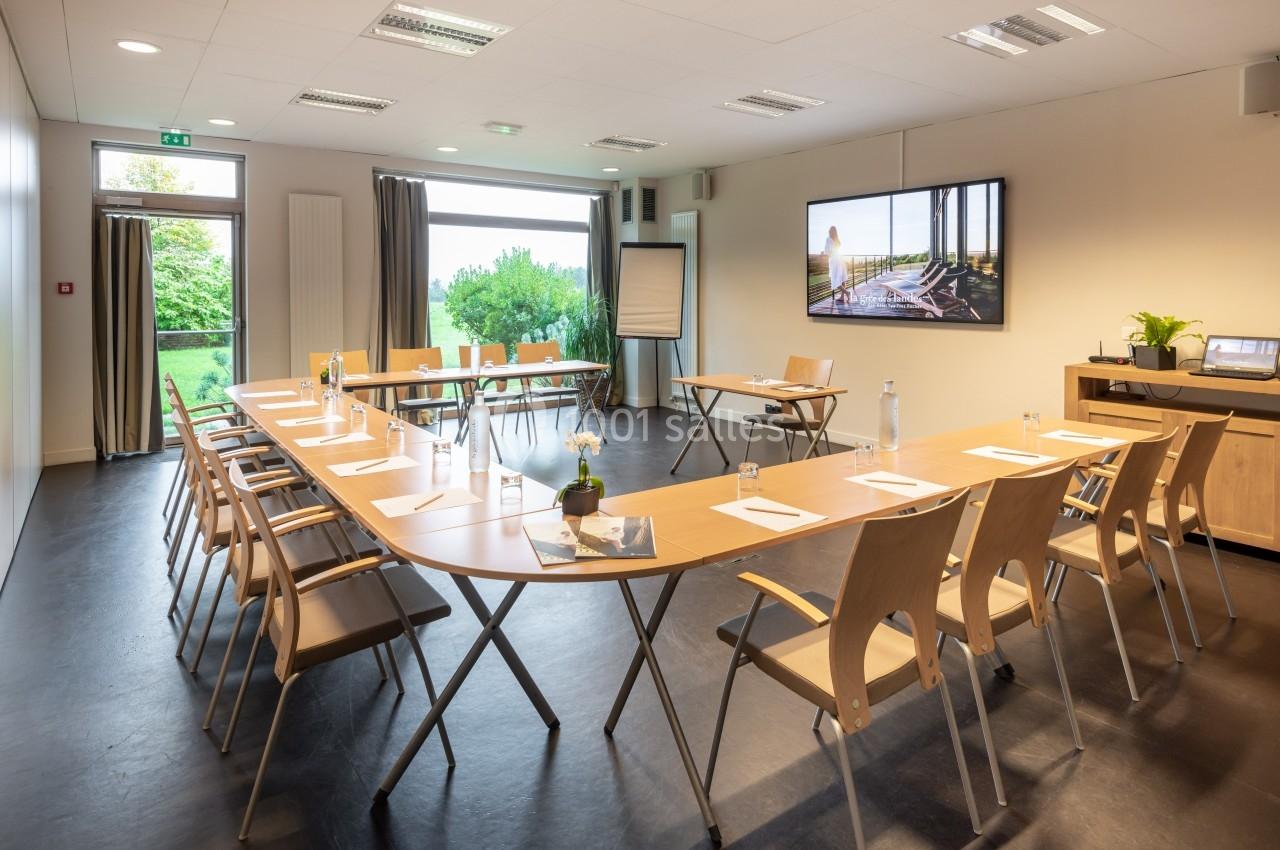 Salle de réunion lumineuse avec tables en U, chaises en bois, écran mural, paperboard et vue sur un jardin.