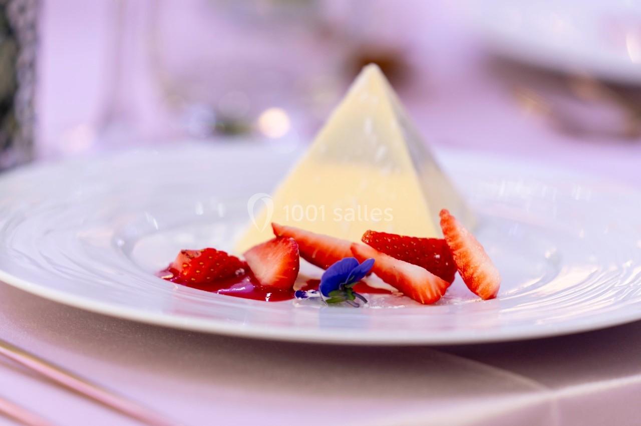 Dessert pyramidal en fromage blanc accompagné de fraises tranchées et d'une fleur comestible sur une assiette blanche.