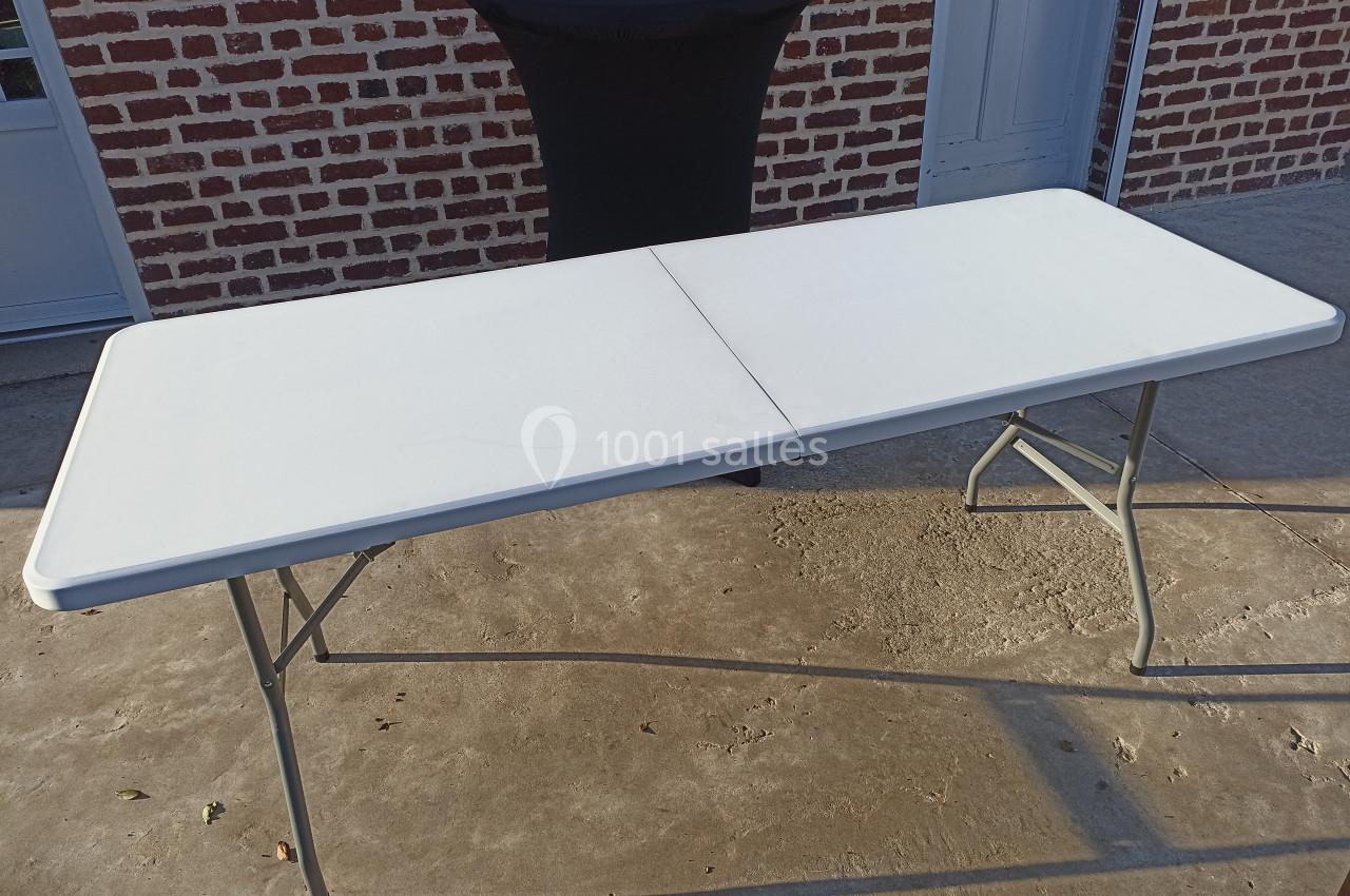 Table pliante blanche en plastique avec pieds métalliques, posée sur une surface en béton devant un mur en briques.