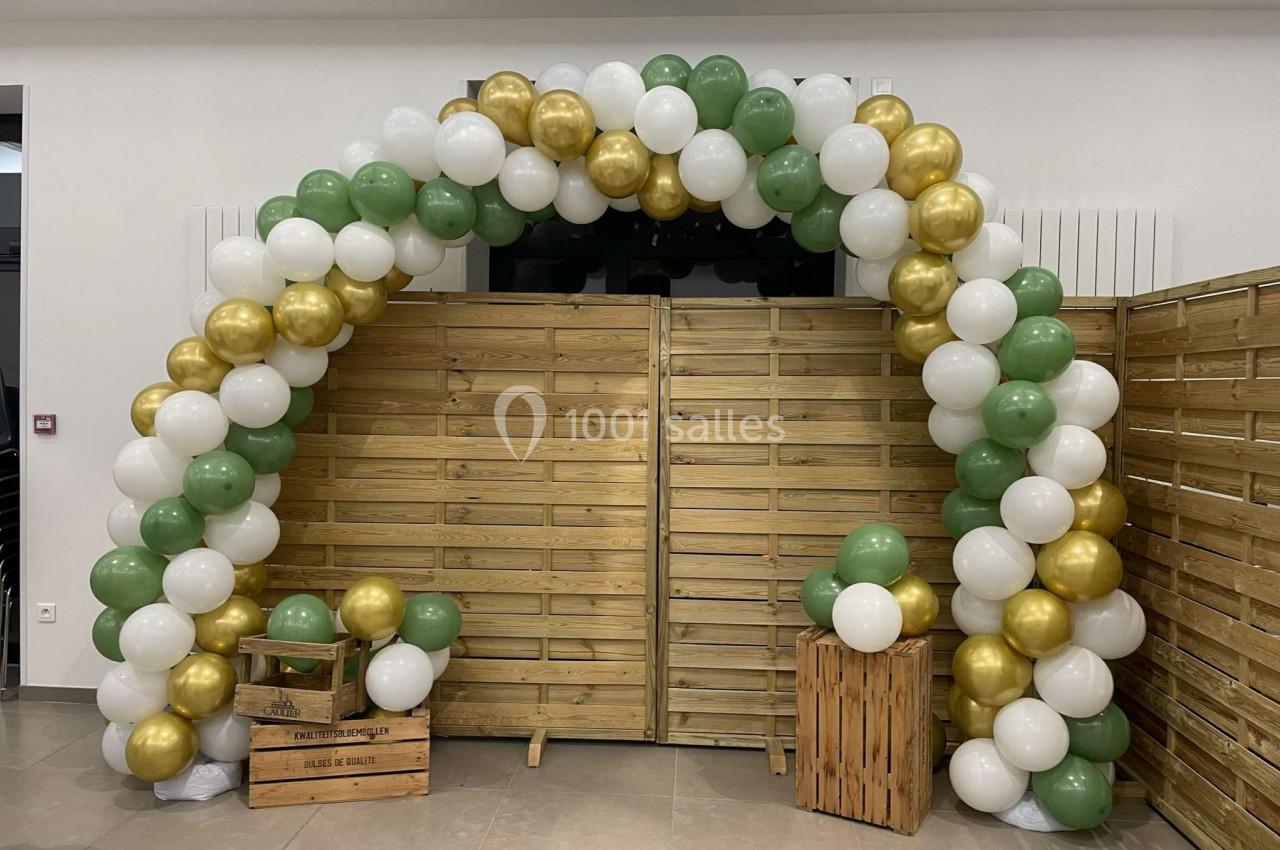 Arche décorative en ballons blancs, verts et dorés devant une cloison en bois, avec des caisses en bois au sol.