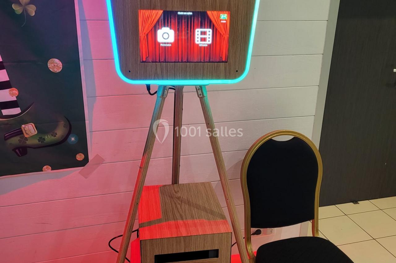 Borne photo interactive en bois avec écran tactile, posée sur un trépied, accompagnée d'une chaise noire.