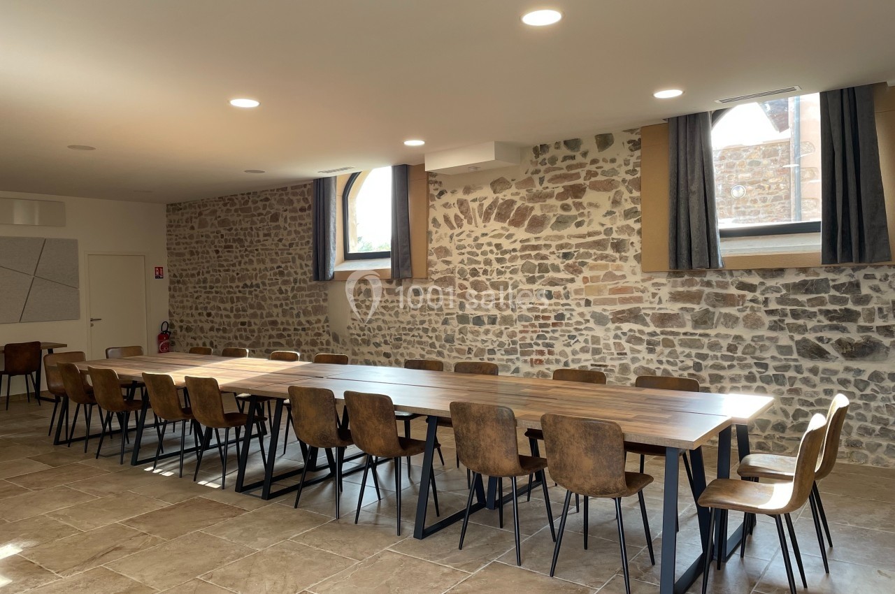 Salle lumineuse avec murs en pierre, grande table en bois entourée de chaises, sol carrelé et fenêtres avec rideaux.