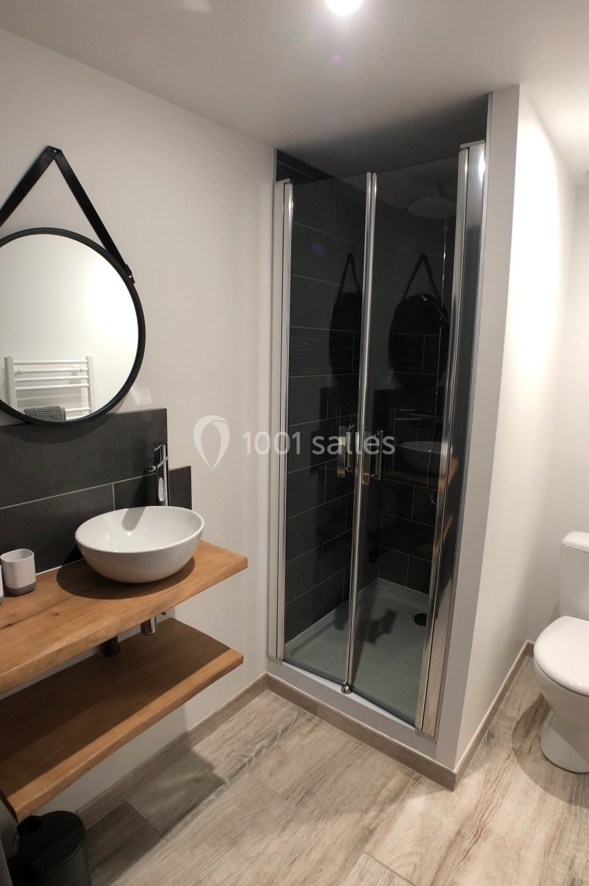 Salle de bain moderne avec douche vitrée, lavabo sur plan en bois, miroir rond et toilettes blanches.