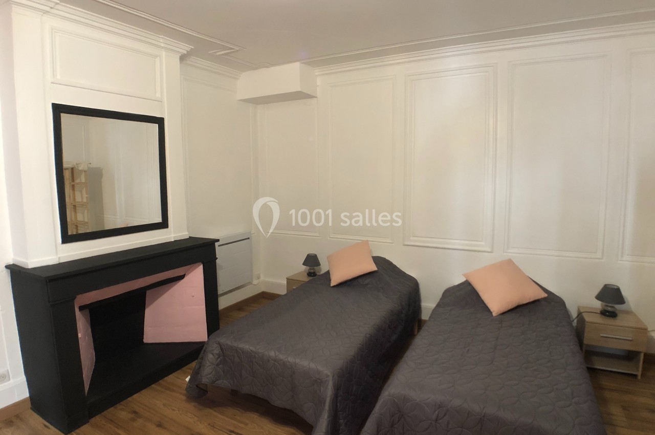 Deux lits simples avec couvre-lits gris, une cheminée noire, un miroir et des murs blancs dans une chambre lumineuse.