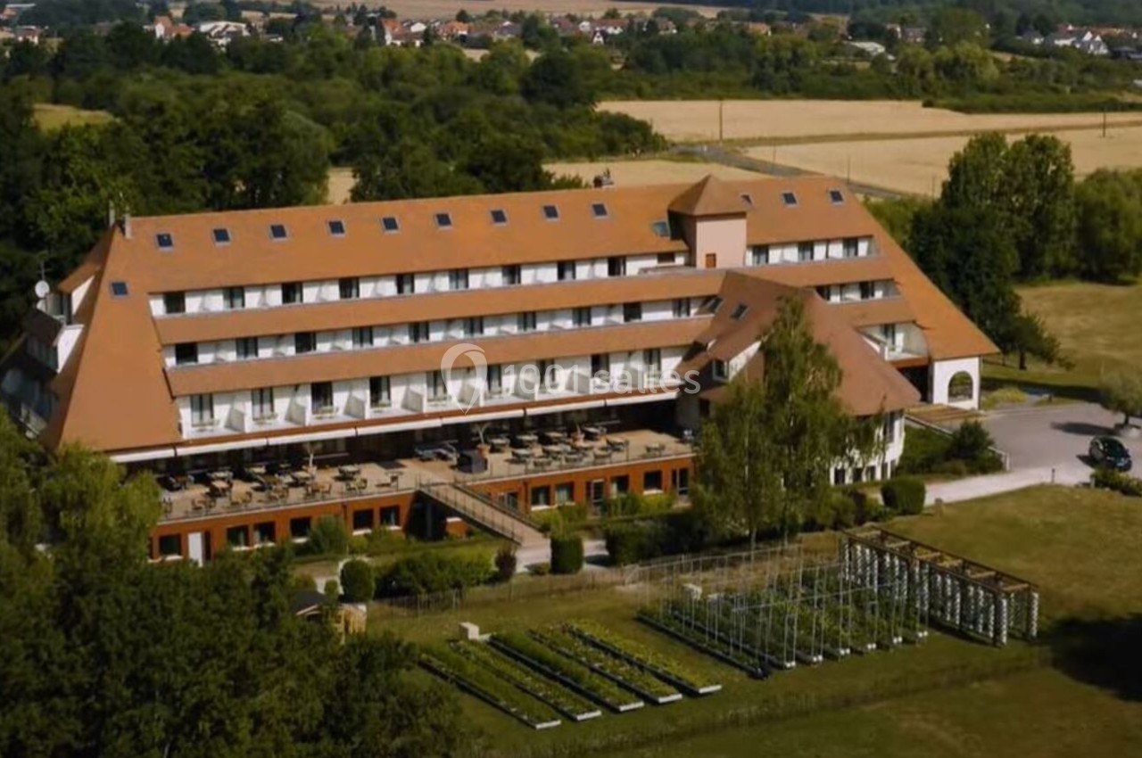 Vue aérienne d'un grand bâtiment avec toit en pente, entouré de verdure et de champs, avec un jardin aménagé au premier plan.
