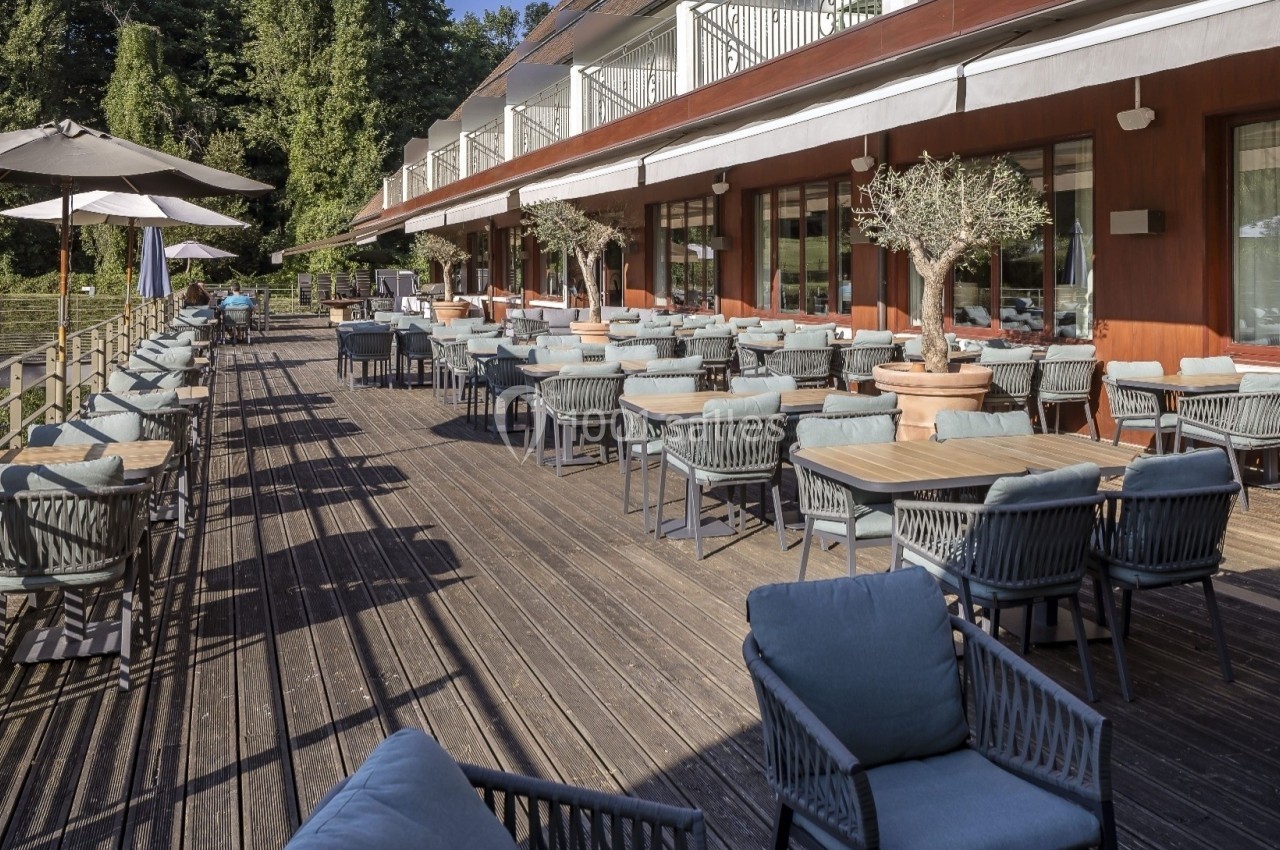 Terrasse en bois avec tables, chaises et parasols, bordée de plantes en pots, devant un bâtiment moderne.